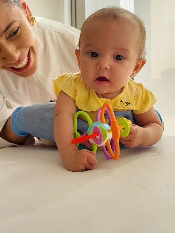 Viviane Vasconcelos sorri ao lado de um bebê de bruços que brinca com um mordedor colorido