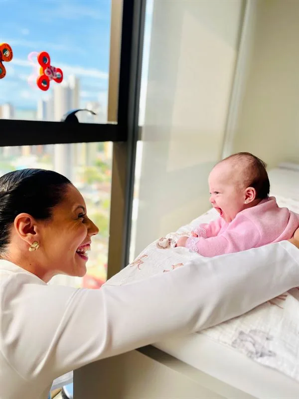 Viviane Vasconcelos sorri para uma bebê de casaquinho rosa, de bruços perto da janela