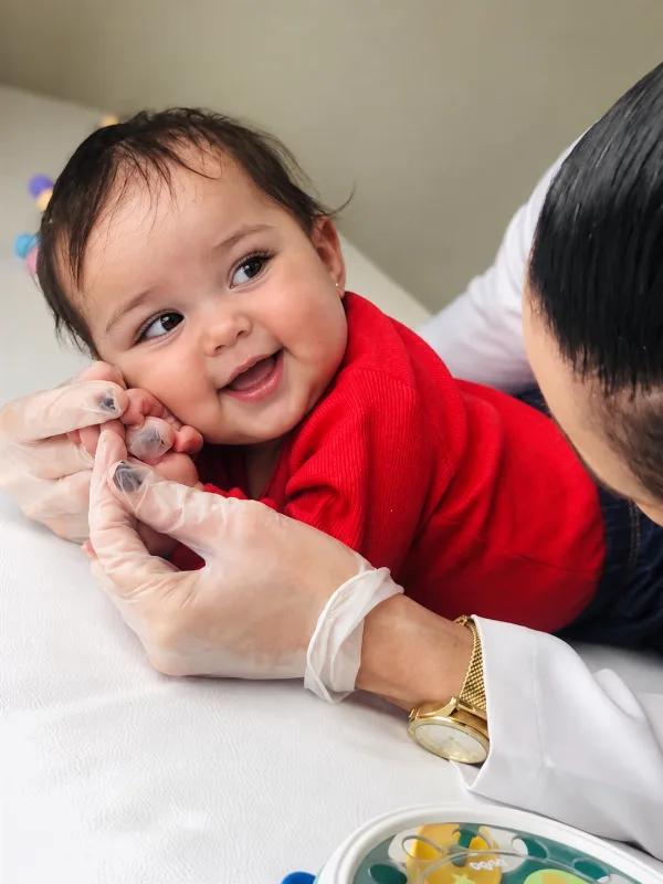 Bebê sorrindo de lado, deitado de bruços, enquanto a fisioterapeuta apoia o rostinho com as mãos